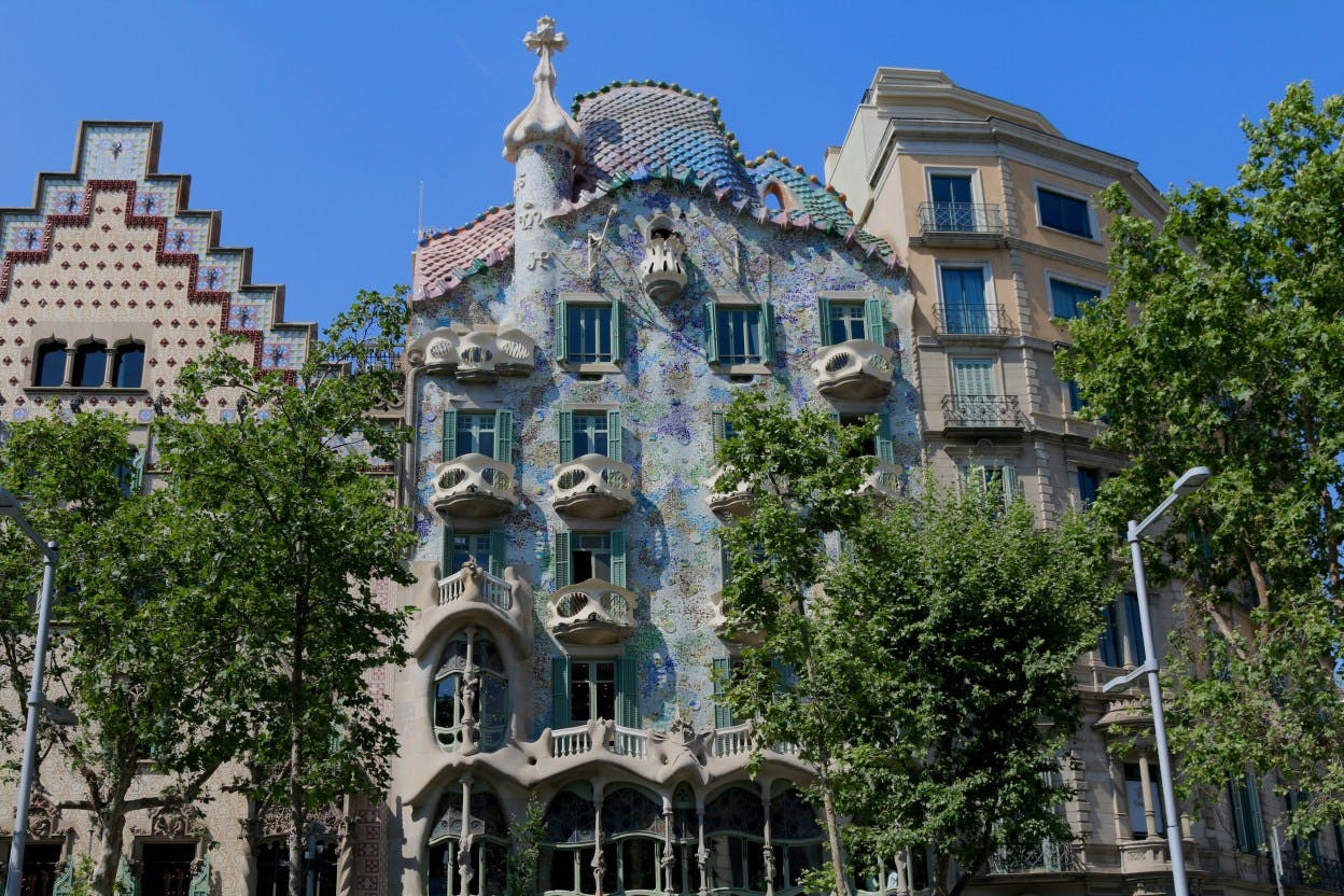 A blue and ornate Gaudi-designed building with bubble-like balconies on Passeig de Gràcia in Barcelona, Spain. The street is lined with trees, and the Gaudi masterpiece, the exterior covered in a material and texture that mimics fish scales, stands out among the other buildings. 