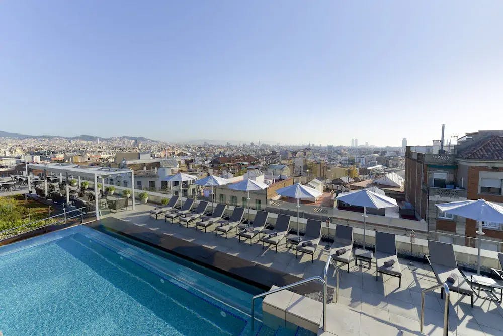 The rooftop pool of the InterContinental Barcelona, an IHG Hotel with beach chairs and sun umbrellas.  Taken in the Poble Sec neighborhood, the hilly area provides panoramic views across the Barcelona skyline and mountains beyond. 

