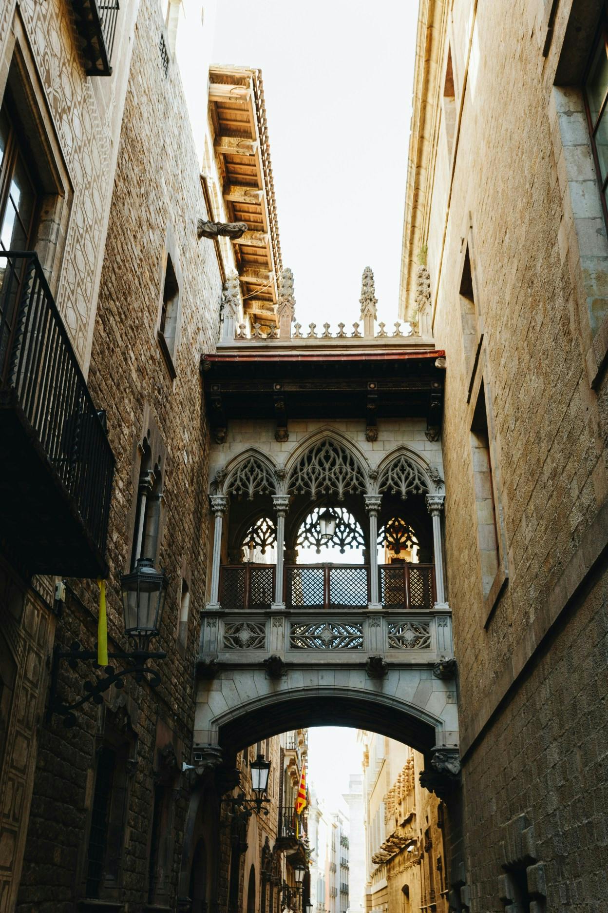 Between two stone buildings, a gothic and medieval bridge passageway connects the two. There are window openings to look down at the alley below. Taken in the Gothic Quarter of Barcelona, Spain. 