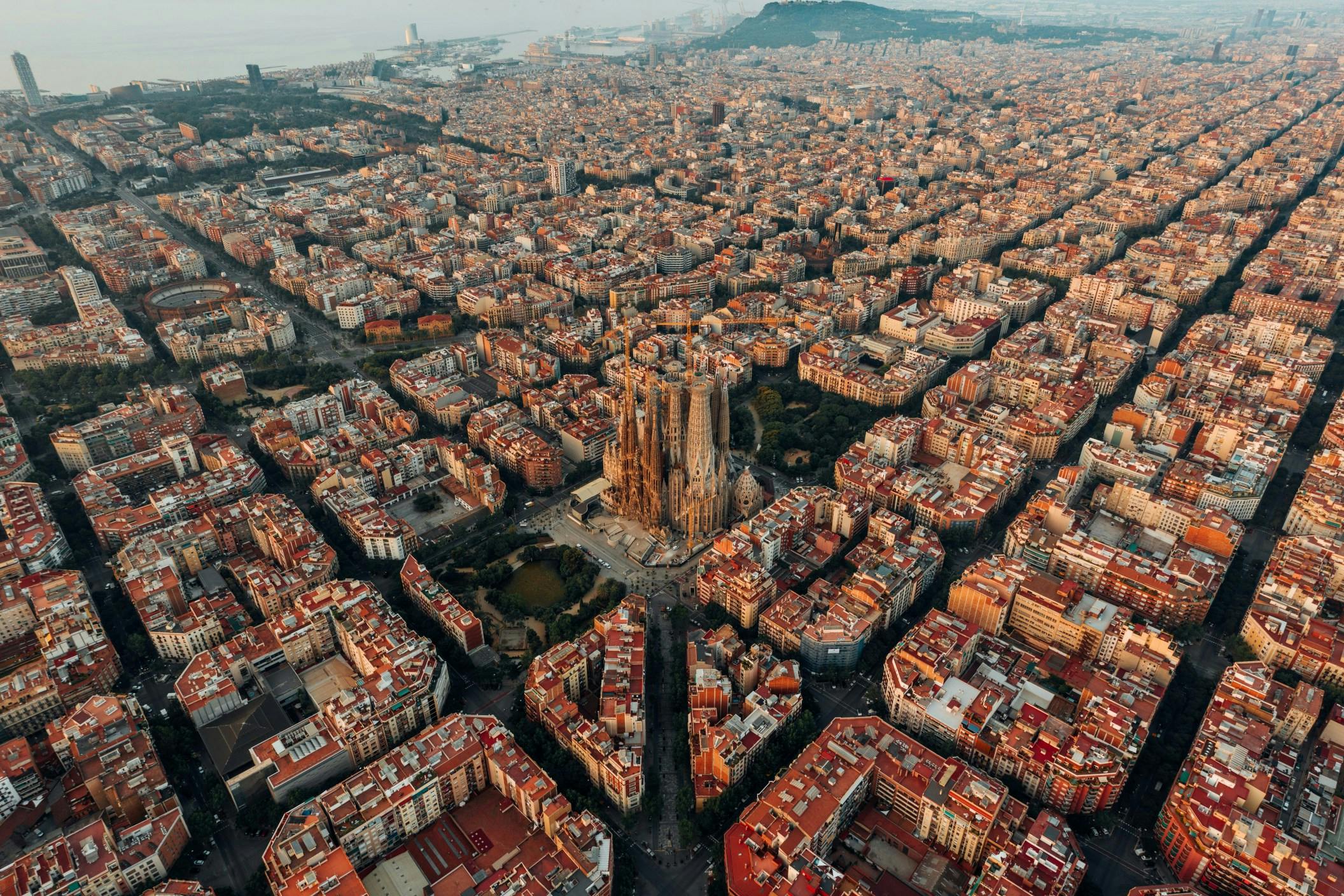 A bird's eye view of the uniform city blocks of Barcelona, Spain. The Sagrada Familia protrudes above the center of the city. The reddish-orange rooftops of the buildings create a glow from the perspective of the image. 