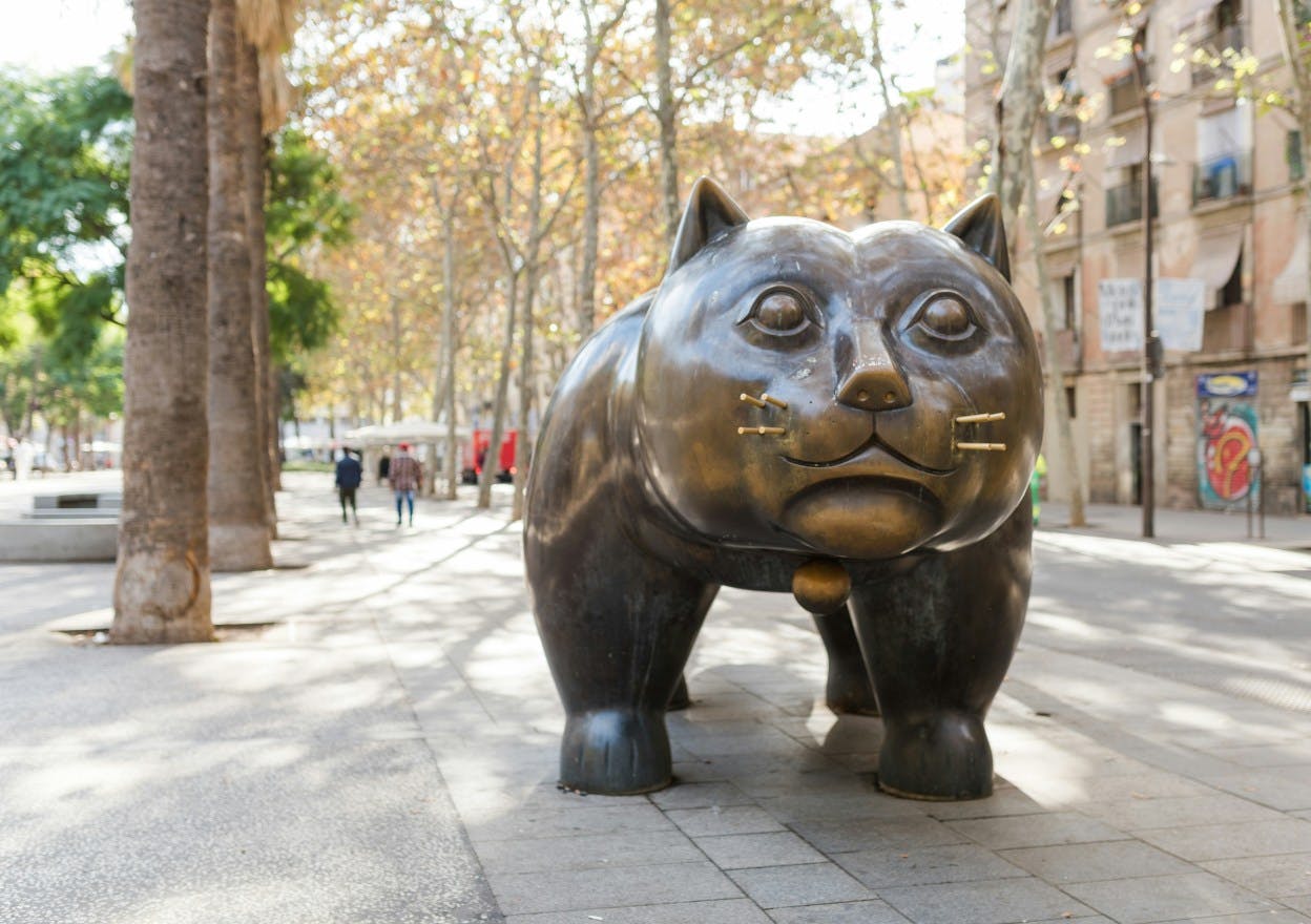 A large metal statue of a cat is positioned in the middle of a city square. Behind the cat, trees and other greenery fill in the square in the El Raval neighborhood of Barcelona. 